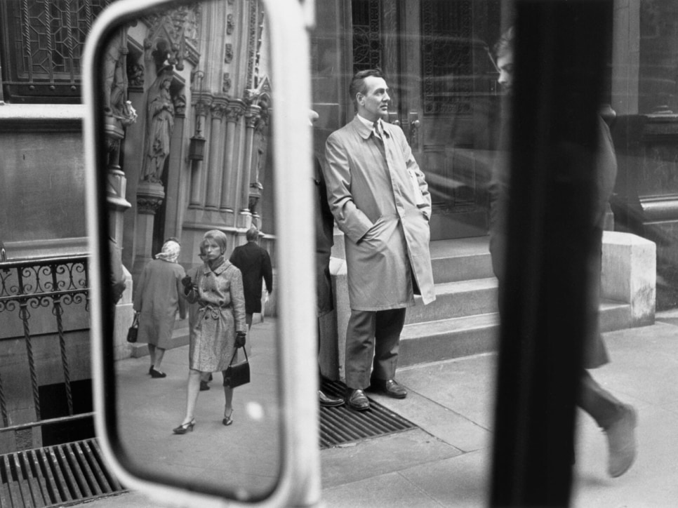 black and white photo of a street taken from a car with the rear view mirror reflecting the other side of the sidewalk, 5 pedestrians
