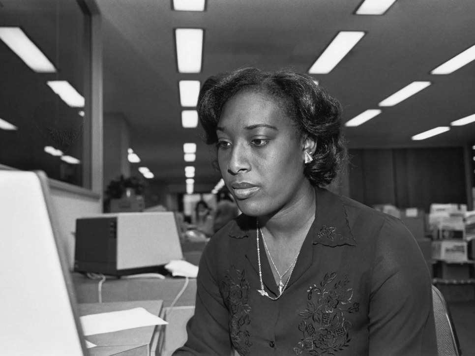 Black and white photograph of a woman sitting in front of a computer in an office, circa 1986