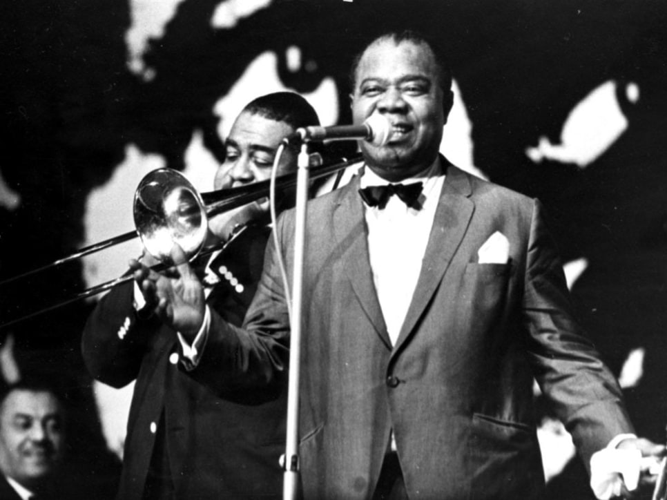 Louis Armstrong at the microphone with a trombone player behind him