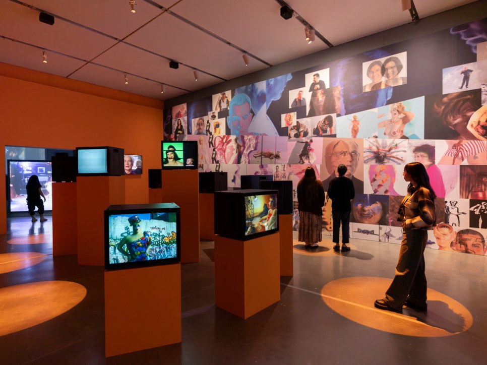 Museum exhibition space with multiple video monitors and a wall filled with various stills of video portraits