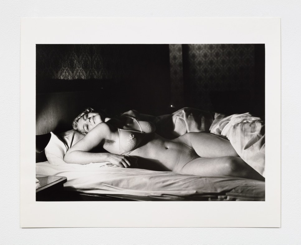 Black and white photographic print of a nude woman laying in bed wearing a bra
