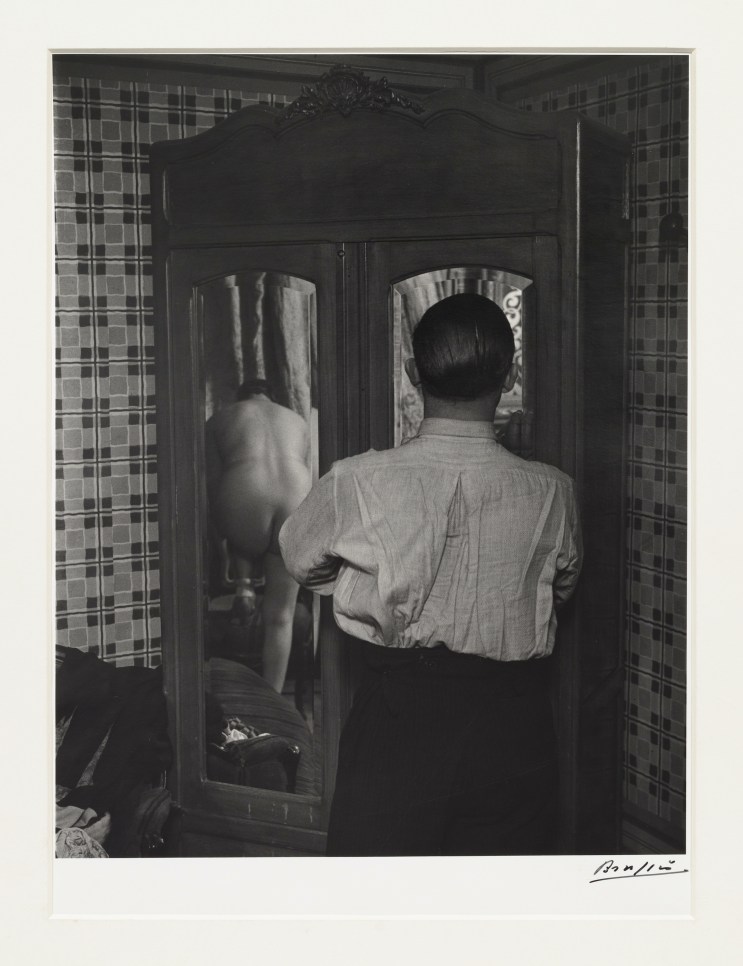 Black and white photographic by Brassaï featuring the behind view of a man looking at the reflection of a woman changing through a mirror
