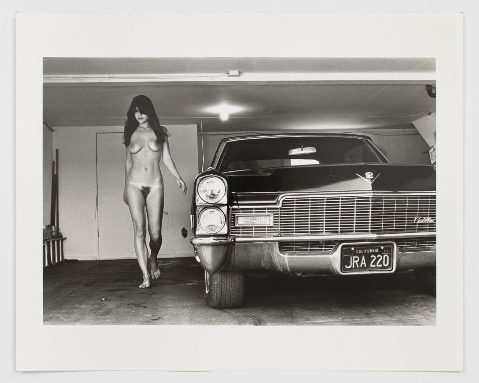 Black and white photographic by Helmut Newton featuring a nude woman posing behind a car in a garage