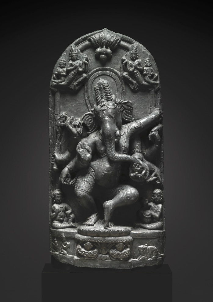 Watched by two cloud-borne celestial garland bearers, eight-armed elephant-headed Ganesha dances in a lively attitude on a lotus flanked by his recumbent rat vehicle and a kneeling donor figure.